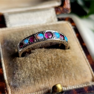 Könnte beinhalten: Ein silberner Ring mit abwechselnd runden Edelsteinen aus Rubin und Opal. Der Ring ist in einem dekorativen Band gefasst und wird in einer Vintage-Schmuckschatulle präsentiert. Die Edelsteine haben einen Durchmesser von ca. 3 mm.