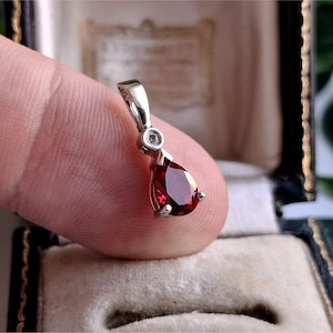 May include: A silver-toned pendant featuring a pear-shaped, deep red faceted gemstone at the base. Above the main stone, a small, clear, round faceted stone is set into the metal bail, which connects to a polished silver-toned loop for a chain.