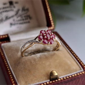 Può includere: Un anello d'oro con un gruppo di pietre preziose rosse, probabilmente rubini, disposte a forma di fiore. L'anello è presentato in un portagioie rosso con interno color crema. La fascia dell'anello ha piccole pietre chiare.