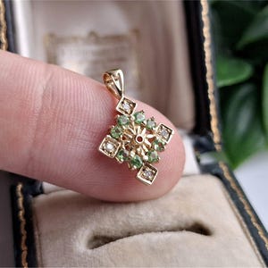 May include: A gold pendant featuring a square-shaped design with a central starburst pattern, surrounded by small round green gemstones and four clear accent stones set at the corners of the frame, complete with a small triangular bail at the top.