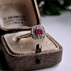 May include: A vintage ring featuring a square, deep red gemstone at its centre, encircled by smaller, clear stones. The ring has a gold band and is presented in a brown, antique jewellery box with a beige interior.