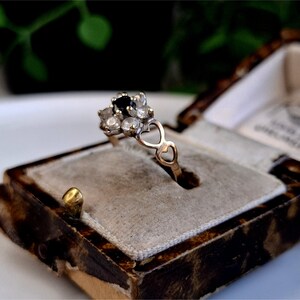 Op de afbeelding: Een vintage gouden ring met een cluster van kleine, heldere stenen en een enkele donkere steen in het midden. De ring heeft een hartvormig ontwerp en wordt gepresenteerd in een decoratieve, antieke sieradendoos.