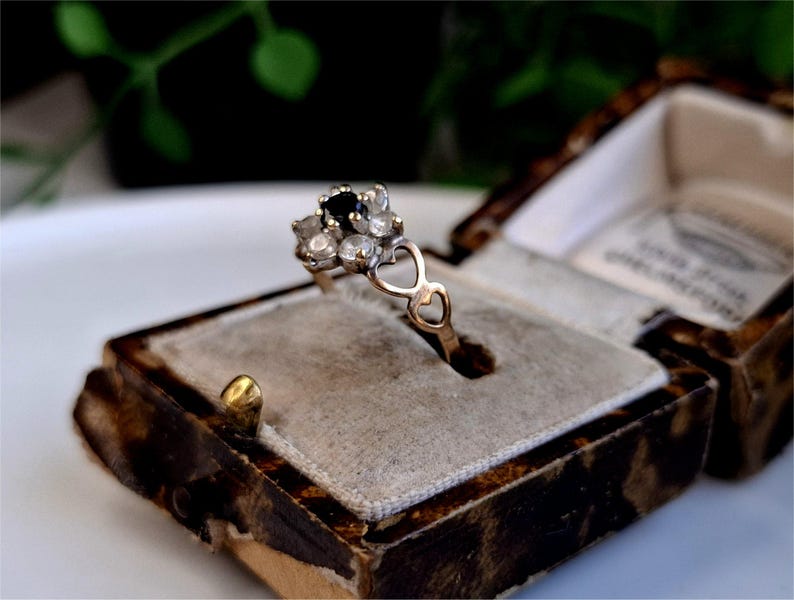 Op de afbeelding: Een vintage gouden ring met een cluster van kleine, heldere stenen en een enkele donkere steen in het midden. De ring heeft een hartvormig ontwerp en wordt gepresenteerd in een decoratieve, antieke sieradendoos.