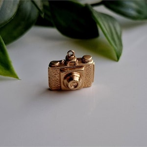 May include: A small, gold-coloured camera charm with intricate detailing. The charm features a classic camera design with a lens, buttons, and a top ring for attachment. The charm is set against a white surface with green leaves in the background.