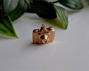 May include: A small, gold-colored camera charm with intricate detailing. The charm features a classic camera design with a lens, buttons, and a top ring for attachment. The charm is set against a white surface with green leaves in the background.