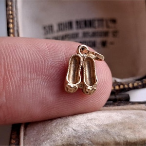 Pode incluir: Um pequeno berloque dourado em forma de par de sapatilhas de ballet. O berloque é detalhado com pequenos laços nos dedos e uma argola para prender a uma pulseira ou colar. O berloque é segurado entre os dedos.