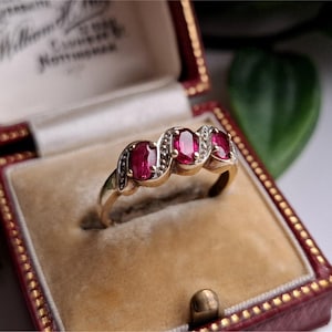 Pode incluir: Um anel de ouro com três pedras preciosas vermelhas ovais, cada uma rodeada por pedras claras menores. O anel é exibido em uma caixa de joias vintage vermelha e bege. A caixa tem detalhes dourados e um interior creme.