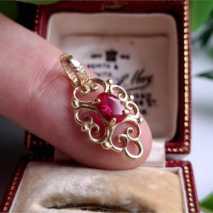 Peut inclure: Un pendentif en or avec une pierre précieuse rouge ovale sertie dans un motif filigrané orné. Le pendentif est présenté dans un écrin rouge avec un intérieur crème.