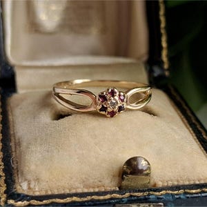 May include: A gold ring with a floral design, featuring small red gemstones and a central clear stone. The ring is displayed inside a vintage blue jewelry box with a gold trim and a beige interior.
