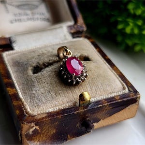 May include: A red ruby gemstone pendant with a gold setting and a halo of smaller diamonds. The pendant is in a velvet-lined jewelry box.