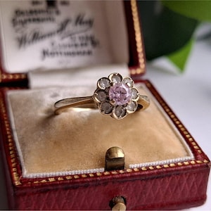 Puede incluir: Un anillo de oro con un diseño en forma de flor, con una piedra central rosa rodeada de piedras claras más pequeñas. El anillo se muestra en un joyero rojo con interior color crema.