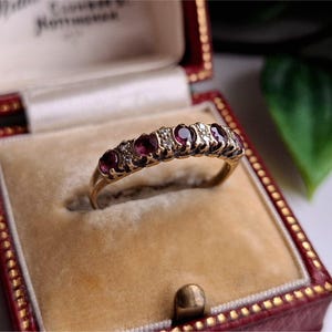 Könnte beinhalten: Ein goldener Ring mit fünf oval geschliffenen, tiefroten Edelsteinen und kleinen, klaren Akzentsteinen. Der Ring wird in einer rot-goldenen Schmuckschatulle mit cremefarbenem Innenraum präsentiert.