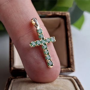 May include: A gold-coloured cross pendant set with emerald-green gemstones. The cross is held against a neutral background, highlighting the design and the vibrant green of the stones. The pendant is a piece of jewellery.