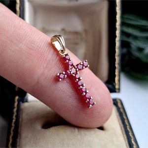 May include: A gold cross pendant featuring multiple small, round, deep red rubies set along the entire length of the cross. The pendant includes a gold bail at the top for attachment to a chain, showcasing a classic design with vibrant red stones.