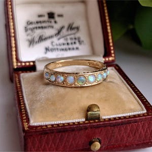 Puede incluir: Un anillo de banda de oro con una hilera de pequeños ópalos redondos e iridiscentes engastados en un marco de metal decorativo. El anillo se muestra dentro de un joyero rectangular de color rojo oscuro forrado de terciopelo con texto.