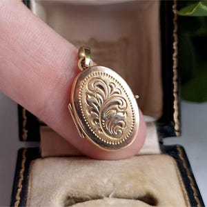 May include: A gold oval locket with an ornate floral design. The locket is held between fingers and is open, revealing a small space inside. The locket has a textured border and a loop for a chain. The background is a black box with a beige interior.