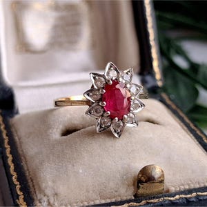 May include: A gold ring featuring a central oval-cut red gemstone surrounded by a halo of twelve small, clear, round-cut stones set in a star-shaped silver-toned metal frame. The ring is displayed in a dark, velvet-lined jewelry box.