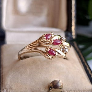 Könnte beinhalten: Ein goldener Ring mit floralem Design, besetzt mit oval geschliffenen Rubin-Edelsteinen und kleinen Diamanten. Der Ring wird in einer Vintage-Schmuckschatulle mit beigem Samtinterieur präsentiert. Das Design des Rings umfasst blattartige Elemente.