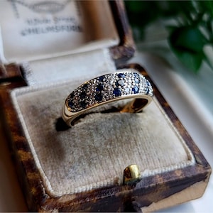 May include: A gold band ring with a row of alternating blue and white gemstones. The ring is in a brown velvet ring box.