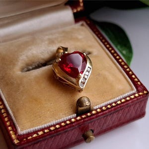 Può includere: Un ciondolo a forma di cuore con una grande pietra preziosa rossa sfaccettata incastonata in metallo dorato. Il ciondolo presenta una piccola fila verticale di tre pietre d'accento chiare e rotonde lungo un lato della cornice a cuore.