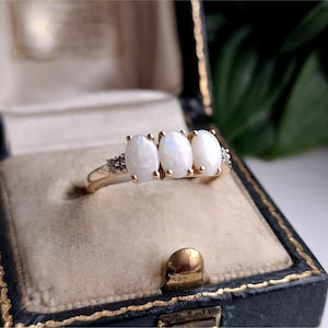 May include: A gold ring featuring three oval-shaped white opals set in a row, accented by small clear stones on each side of the band. The ring is displayed inside a dark blue, velvet-lined jewellery box with gold trim along the edges.