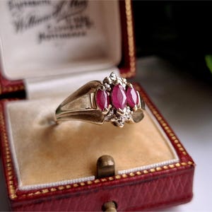 May include: A gold ring featuring three oval, deep red gemstones, set with smaller accent stones. The ring is presented in a red jewellery box with a cream interior.