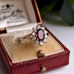 May include: A gold ring featuring a central oval-cut, deep red gemstone, encircled by a halo of smaller, clear gemstones. The ring is presented in a red jewellery box with a cream-coloured interior.