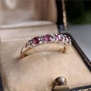 May include: A gold and silver ring featuring five round, pink gemstones and small white stones. The ring is presented in a vintage jewellery box with a beige interior and a dark blue exterior.