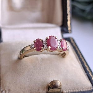 Op de afbeelding: Een gouden ring met drie ovaal geslepen rode robijnen op een rij. De middelste robijn is iets groter dan de twee robijnen aan de zijkanten. Kleine, heldere accentstenen zijn tussen de robijnen in de gepolijste gouden band gezet.