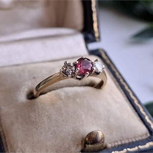May include: A gold ring featuring a central round-cut pink gemstone flanked by two smaller clear round-cut stones. The ring is set in a metal band and presented inside a dark blue velvet-lined jewellery box with a cream-coloured interior cushion.