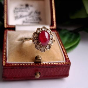 Puede incluir: Un anillo de oro con una gema roja oscura ovalada en el centro, rodeada de piedras claras más pequeñas. El anillo se exhibe en un joyero rojo forrado de terciopelo con detalles dorados.