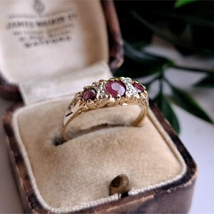 May include: A gold ring featuring three round red gemstones set in a row, accented by small clear stones between them. The band has a decorative engraved pattern along the sides, and the ring is presented in a vintage-style jewelry box.