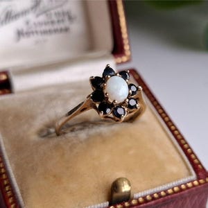 Op de afbeelding: Een gouden ring met een ovale opaal edelsteen in het midden, omringd door donkerblauwe saffieren. De ring, met een bloemmotief, wordt gepresenteerd in een rode fluwelen sieradendoos.