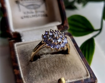 May include: A gold ring with a cluster of purple amethyst stones set in a flower shape. The ring is in a brown velvet ring box.