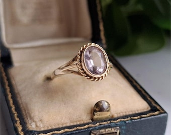 Puede incluir: Un anillo de oro con una piedra preciosa ovalada de color púrpura claro, engastada en un bisel retorcido en forma de cuerda. El anillo se exhibe dentro de un joyero azul vintage con ribete dorado e interior beige.