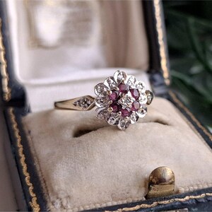 May include: A gold ring featuring a floral-shaped cluster setting with a central diamond surrounded by six round-cut red rubies, further accented by a halo of small diamonds and additional diamonds set into the shoulders of the thin gold band.