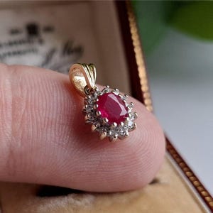 Puede incluir: Un colgante de oro con una piedra preciosa de rubí ovalada en el centro, rodeada de piedras preciosas claras más pequeñas. El colgante se muestra sobre un fondo borroso, destacando el diseño y el color de la joya.