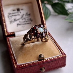 May include: A vintage gold ring with a cluster of red gemstones surrounding a central white opal. The ring is displayed in a red velvet-lined jewelry box, showcasing its intricate design and craftsmanship. The ring's setting is ornate.