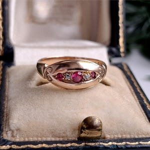 May include: A gold ring featuring a horizontal row of three round red stones alternating with two smaller clear stones, set within a smooth, polished gold band that has decorative engraved scrollwork on the sides of the central setting.