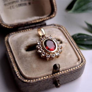 May include: A gold pendant with a large oval, deep red gemstone at its center, surrounded by smaller, clear gemstones. The pendant is displayed in an open, vintage-style jewelry box with a velvet interior.