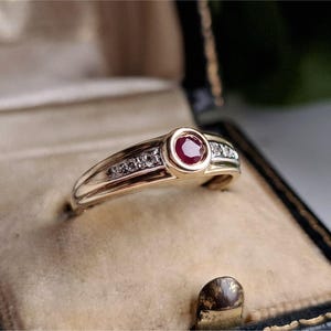 May include: A gold ring featuring a central ruby and small diamonds. The ring is presented in a vintage jewellery box with a beige interior and a black exterior.