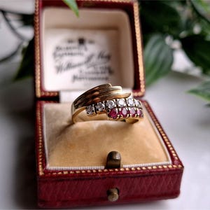 May include: A gold ring set with clear and red gemstones, presented in a red jewellery box with a tan interior. The ring has a layered design with multiple gemstones. The box is open, showcasing the ring and its interior lining.