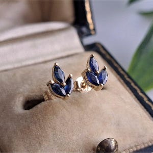 May include: A pair of gold stud earrings with three dark blue marquise-cut gemstones arranged in a leaf-like design. The earrings are displayed in a vintage jewelry box with a beige interior.