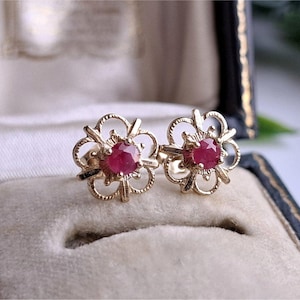 May include: A pair of gold stud earrings featuring a central round-cut red gemstone surrounded by a delicate, openwork filigree metal frame shaped like a stylized flower. The earrings are positioned side by side, showcasing the intricate metalwork.