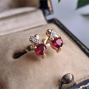 May include: A pair of gold stud earrings, each with a heart-shaped, deep red gemstone. The earrings have a small, curved accent with embedded clear stones. The earrings are presented in a vintage jewellery box.