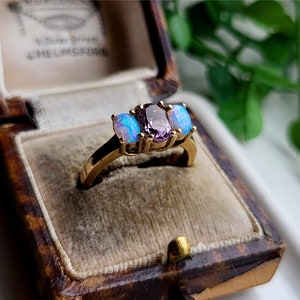 May include: A gold ring with three oval-shaped gemstones. The center stone is a purple amethyst, and the two outer stones are opalescent.