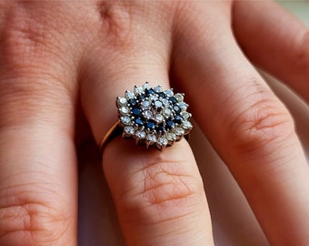 May include: A gold ring featuring a cluster of small, round gemstones. The ring's center has a collection of dark blue stones surrounded by white stones, creating a floral pattern. The ring is set against a blurred background.
