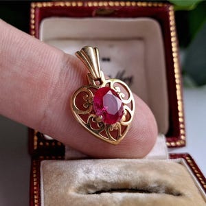 May include: A gold heart-shaped pendant with a deep red, oval gemstone. The pendant has an open filigree design and is presented in a red jewellery box.