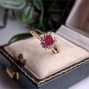 Puede incluir: Un anillo de oro con una piedra central de rubí rojo intenso de corte ovalado rodeada por un halo de pequeñas piedras claras. El anillo tiene un diseño de banda dividida y se muestra dentro de un joyero azul oscuro con interior crema.