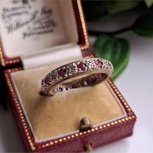 Puede incluir: Un anillo vintage de oro y plata con una banda de piedras preciosas rojas alternas y filigrana decorativa. El anillo se muestra dentro de un joyero rojo con interior color crema.